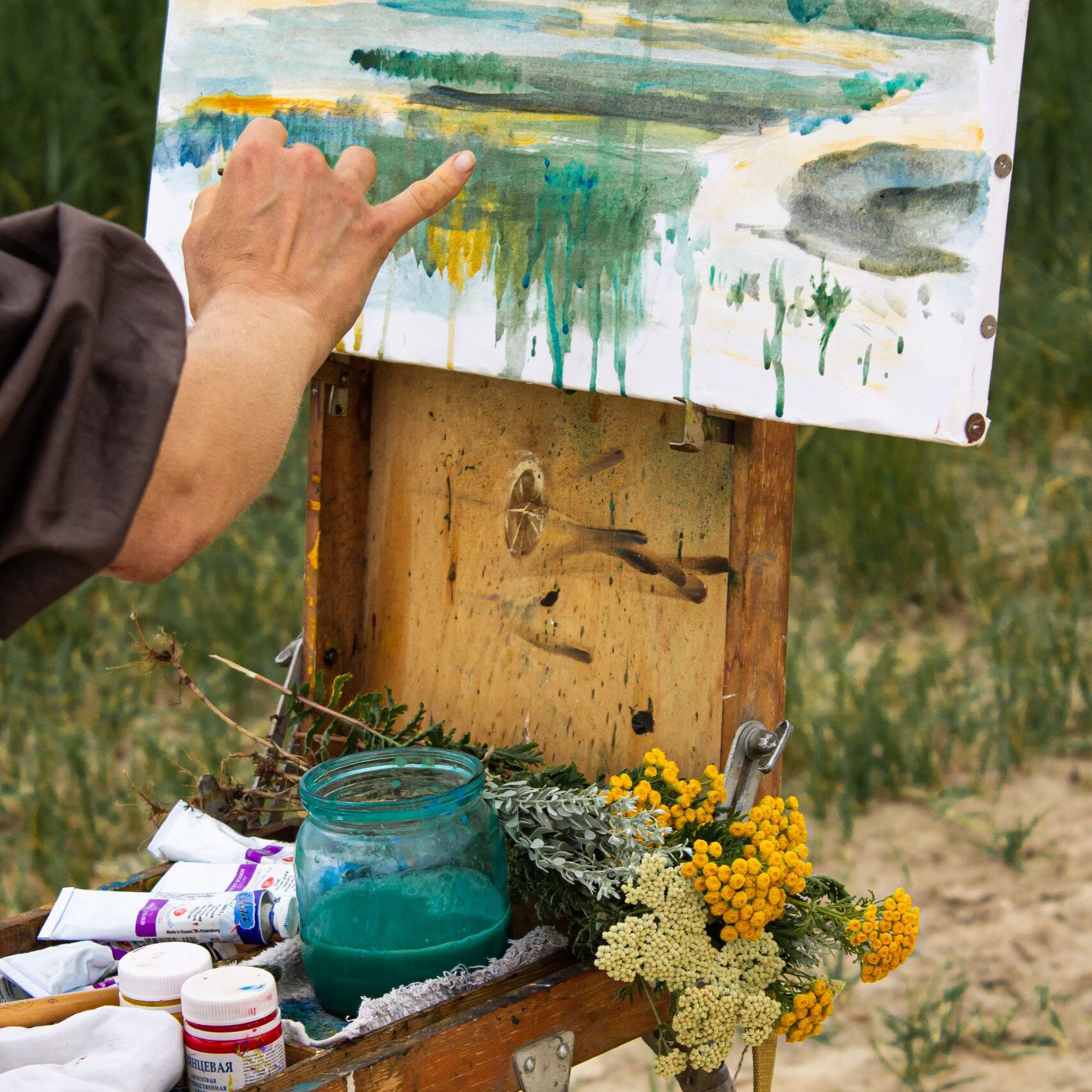 Crop anonymous artist with paintbrush painting scenery of pond in village on easel with paints and flowers